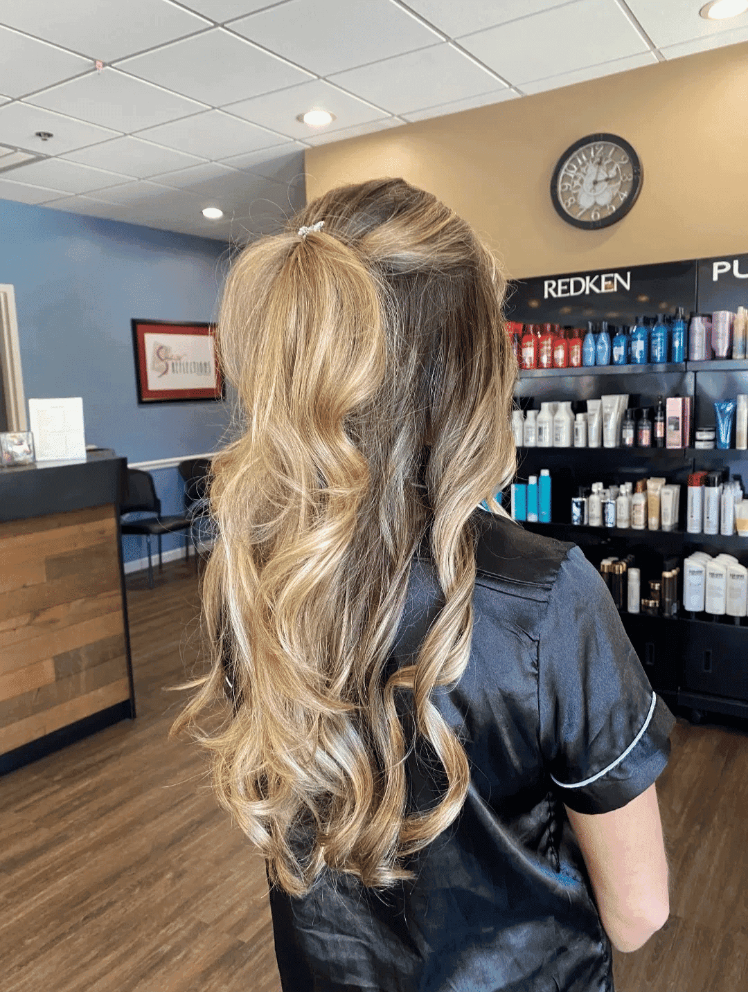 Long, wavy blonde hair styled in a salon, with shelves of hair products in the background.