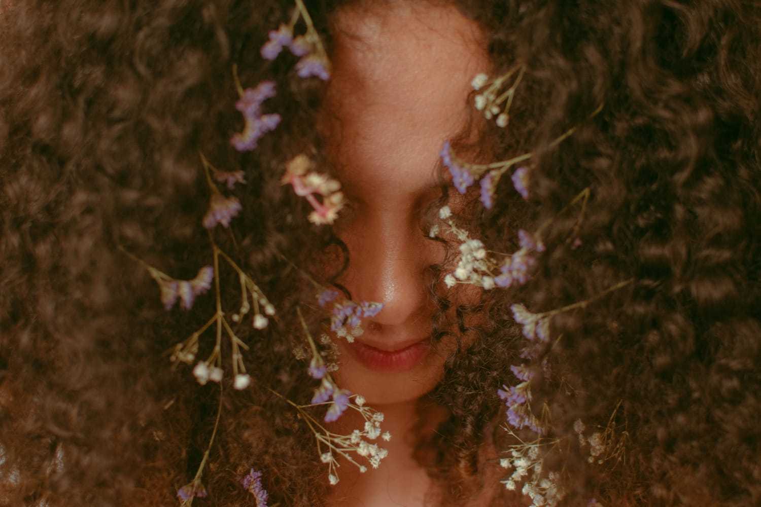 Curly-haired person with delicate purple and white flowers in their hair.