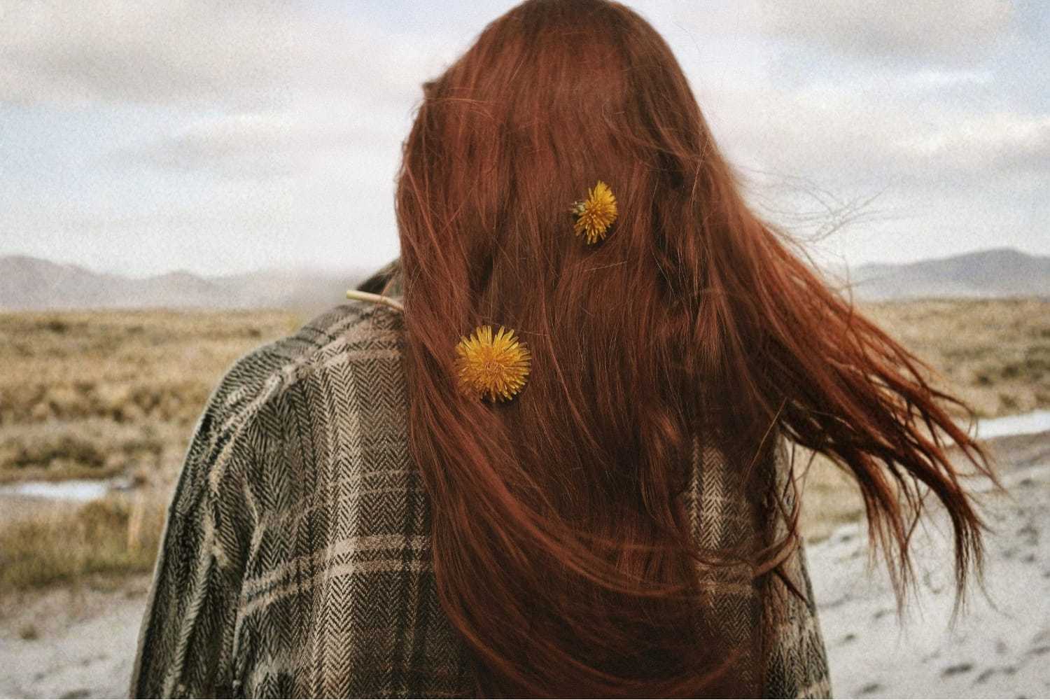 Red-haired person with dandelions in hair stands in windy landscape.