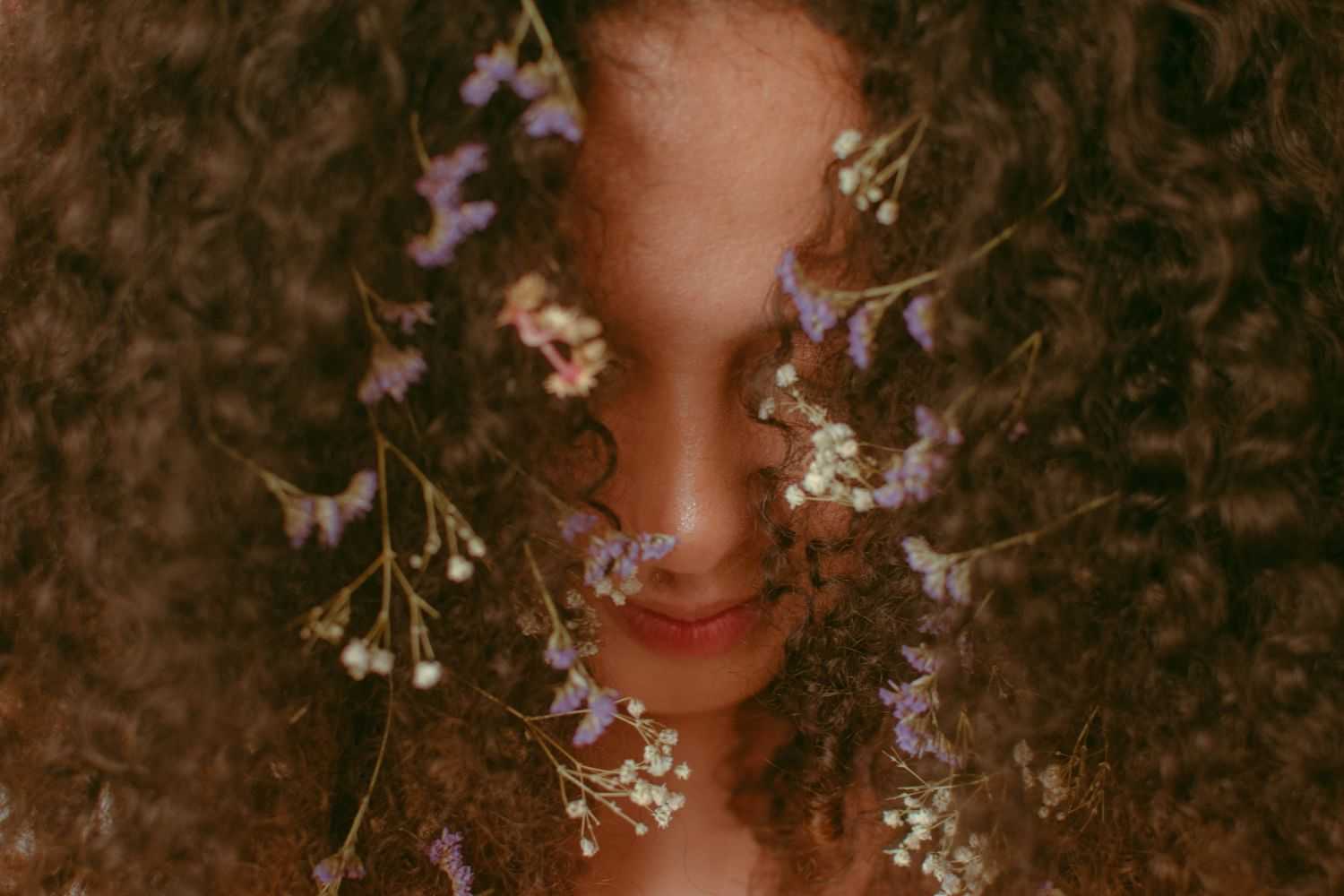 Curly-haired person with delicate purple and white flowers in their hair.